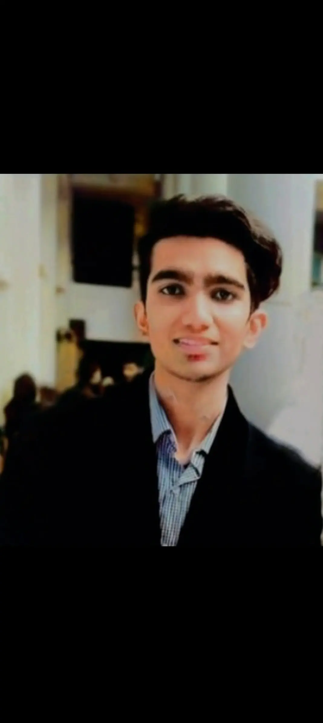 Shehneel with short, dark hair, wearing a dark suit jacket and a light blue shirt, standing indoors with a blurred background.